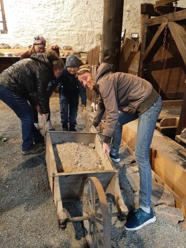 Drei Personen schieben eine alte Schubkarre in einer rustikalen Umgebung.