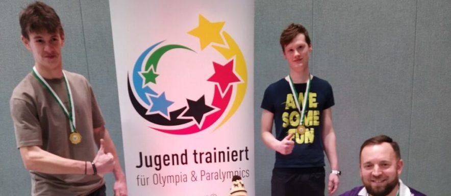Drei Personen mit Medaillen posieren vor einem Banner der Jugendtrainer.
