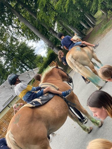 Reiter auf Pferden, die einem Weg folgen, umgeben von grünen Bäumen und Zuschauern.
