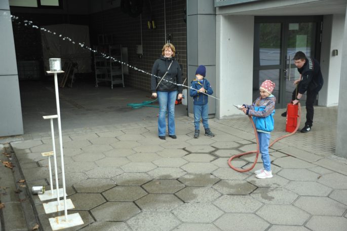 Ein Kind sprüht Wasser mit einem Schlauch, während Erwachsene und ein weiteres Kind zuschauen.