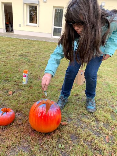 Ein Mädchen mit langen Haaren bemalt einen orangefarbenen Kürbis auf dem Rasen.