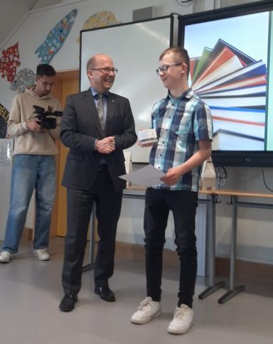 Zwei Personen stehen in einem Raum, eine überreicht einem anderen eine Urkunde.