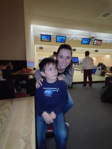 Ein Junge sitzt, während eine Frau hinter ihm steht und lächelt. Bowlingbahn im Hintergrund.