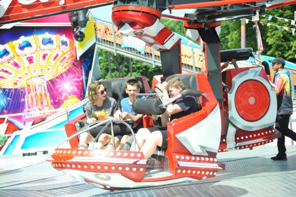 Drei Jugendliche in einem Kettenkarussell, bereit für die Fahrt. Bunte Hintergrundbeleuchtung.