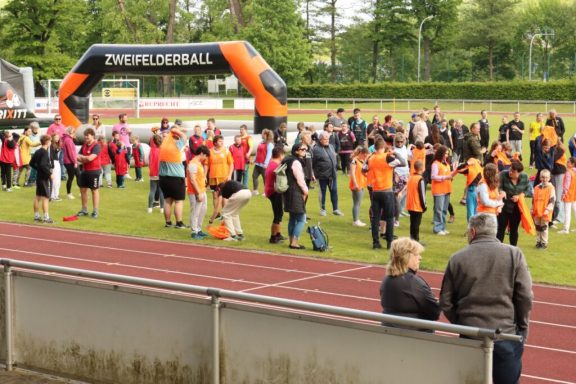 Menschenansammlung auf einem Sportfeld, viele in orangefarbener Kleidung, festivale Atmosphäre.