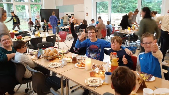 Gruppierung von Kindern und Erwachsenen beim Essen in einem Beisein von Tischen und Stühlen.