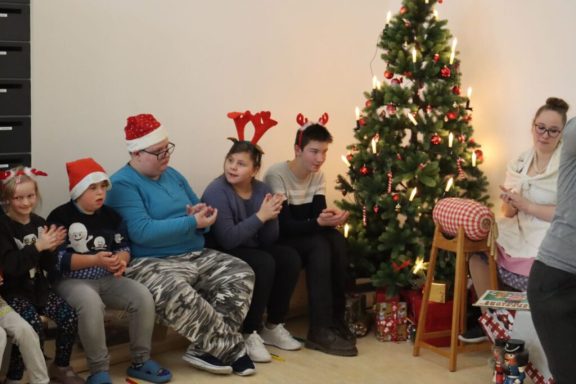Gruppe von Kindern mit Weihnachtsmützen und -hörnern vor einem festlich geschmückten Baum.