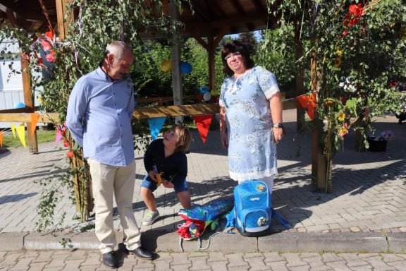 Drei Personen stehen in einem bunten Freizeitumfeld, ein Kind kniet mit Spielzeug.