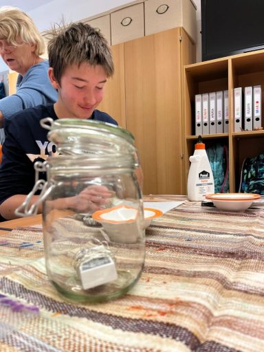 Ein Junge sitzt am Tisch mit einem Glas und Farben, während eine andere Person im Hintergrund steht.