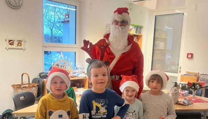 Weihnachtsmann steht mit vier Kindern in Weihnachtsmützen in einem Klassenraum.