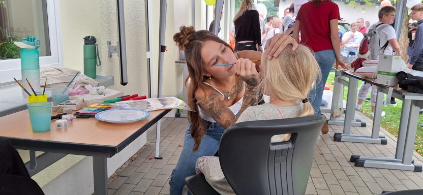 Eine Person schminkt eine andere, während eine bunte Palette und Pinsel auf dem Tisch liegen.