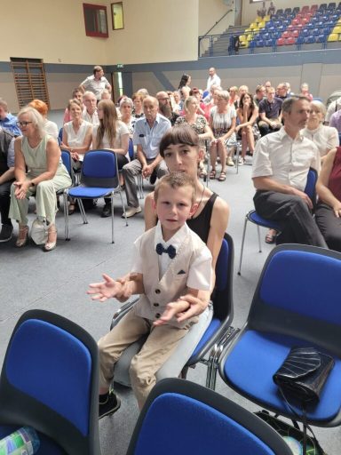 Ein Junge in einem Anzug sitzt mit offenem Ausdruck auf einem Stuhl, vor versammeltem Publikum.
