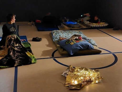 Zwei Personen liegen in Schlafsäcken auf dem Boden, umgeben von Decken und Lichtern.