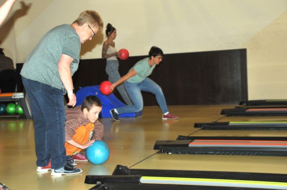 Kinder und Erwachsene spielen zusammen Bowling, einige werfen bunte Bälle auf die Bahnen.