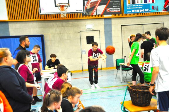 Schüler spielen Basketball in einer Sporthalle, umgeben von Zuschauern und Betreuern.