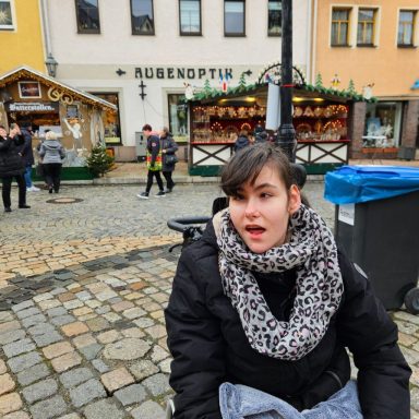 Junge Frau mit einem Schal sitzt auf einem Platz, umgeben von festlich dekorierten Ständen.