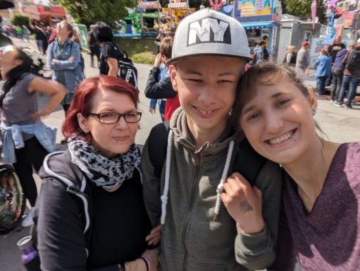 Drei Personen lächeln vor einem bunten Jahrmarkt mit Ständen im Hintergrund.