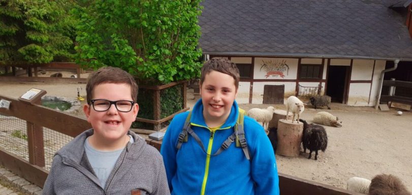 Zwei Kinder lächeln vor einem Bauernhof mit Tieren im Hintergrund.