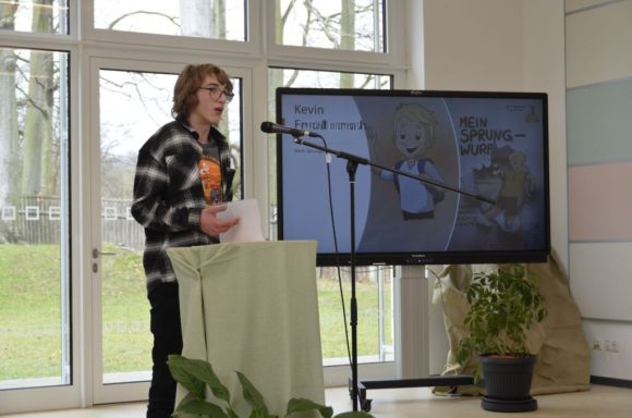 Junger Mann präsentiert vor einem Monitor, umgeben von Pflanzen in einem hellen Raum.