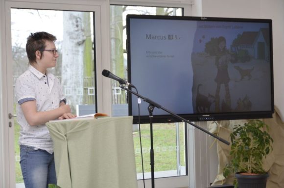 Junger Mann steht an einem Tisch, präsentiert vor einem Bildschirm mit einer Grafik.