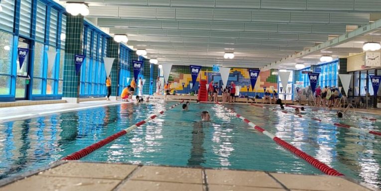 Hallenbad mit Schwimmern, blauen Wänden und einer Wasserrutsche im Hintergrund.