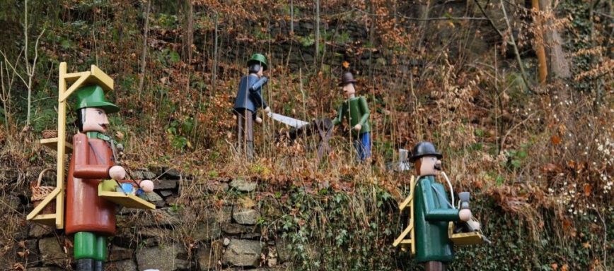 Holzfiguren von Arbeitern in der Landschaft mit Pflanzen und Bäumen im Hintergrund.
