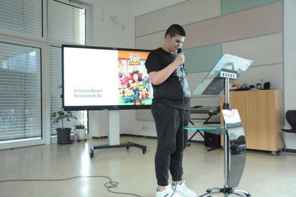 Ein Jugendlicher steht am Mikrofon und hält einen Vortrag vor einer Präsentation.