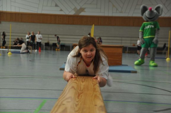 Ein Mädchen zieht an einem Balken, während ein Maskottchen im Hintergrund steht.