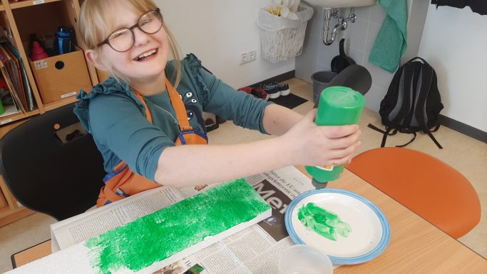 Lachendes Kind in einem blauen Shirt, das grüne Farbe auf einen Teller gießt.