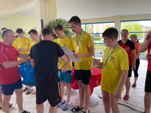 Gruppe junger Schwimmer in gelben T-Shirts erhält Medaillen bei einer Veranstaltung.
