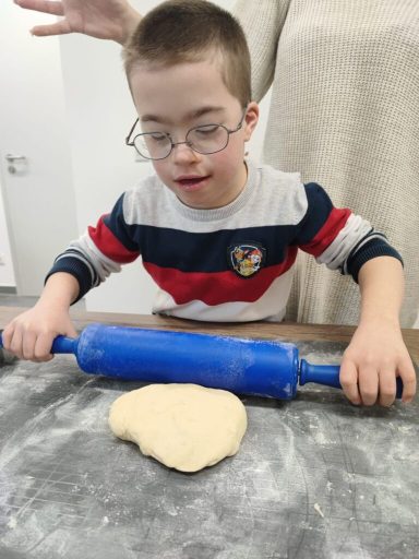 Ein Kind rollt Teig mit einem Nudelholz auf einer bemehlten Arbeitsfläche aus.