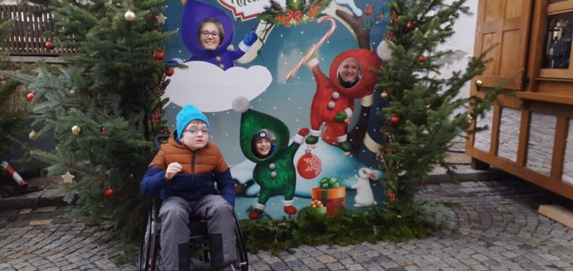 Kind im Rollstuhl vor einer festlich dekorierten Weihnachtskulisse mit anderen Kindern.