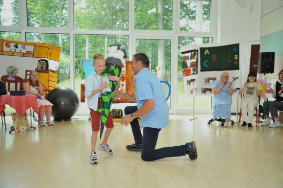 Ein Mann kniet vor einem Jungen in einer bunten Umgebung mit mehreren Erwachsenen im Hintergrund.