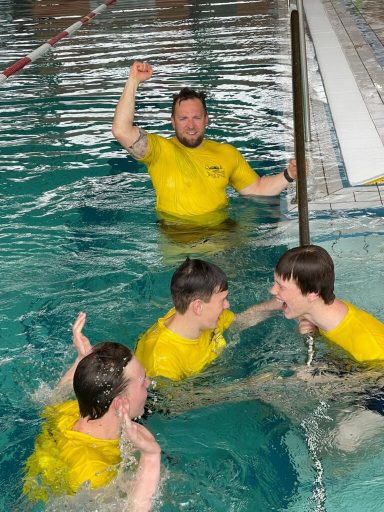 Grupp von vier Jugendlichen in gelben T-Shirts im Wasser, mit einem Schwimmtrainer.
