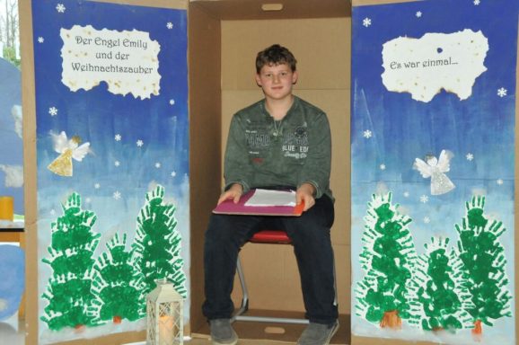 Junge sitzt in einem Kasten mit Weihnachtsdekoration und hält ein Buch auf dem Schoß.