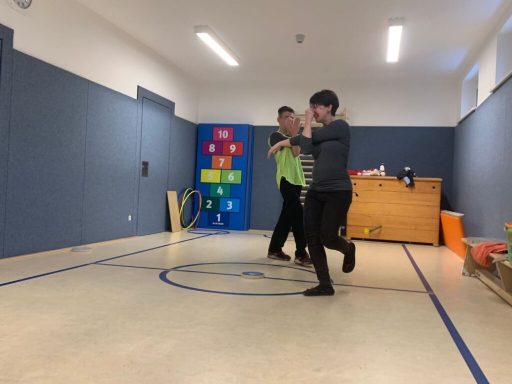 Zwei Personen tanzen in einem Raum mit einer Holzbank und bunten Matten im Hintergrund.