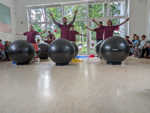 Drei Personen mit großen Bällen in einem Raum, umgeben von Zuschauern, die applaudieren.