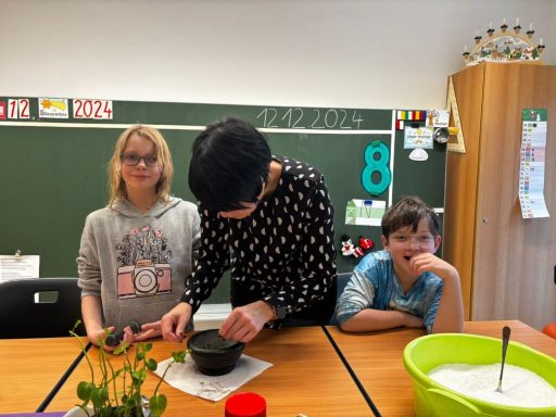 Zwei Kinder stehen an einem Tisch, während ein Erwachsener etwas in einer Schüssel mischt.