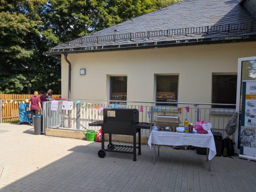 Ein Grillstand mit Tischen und Dekoration vor einem Gebäude, umgeben von Bäumen.