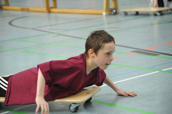 Ein Kind in einem roten Shirt gleitet auf einem Rollbrett über den Hallenboden.