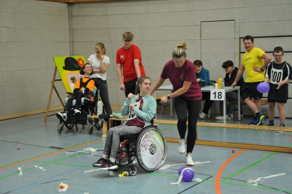 Eine Person im Rollstuhl wird von einer Helferin unterstützt, während andere im Hintergrund aktiv sind.