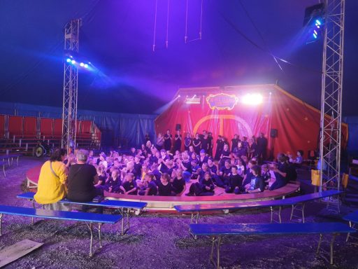 Eine Gruppe von Menschen sitzt im Zirkuszelt, umgeben von bunten Lichtern.