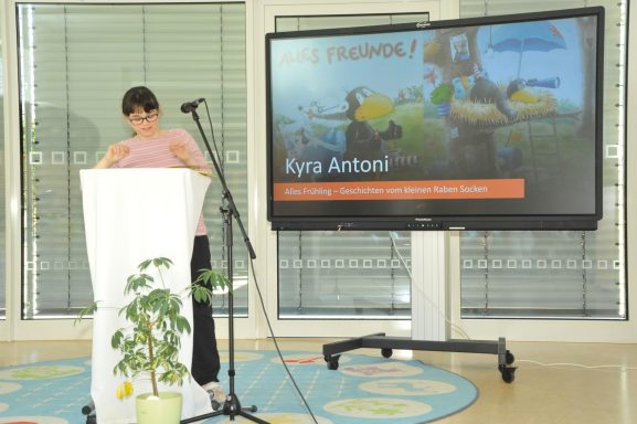 Eine Person steht an einem Podium, während eine Präsentation auf einem Bildschirm gezeigt wird.