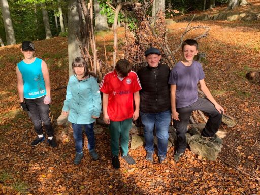 Fünf Personen stehen im Wald auf einem mit Laub bedeckten Erdgrund.