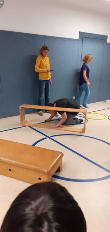 Kinder spielen in einer Turnhalle, einige stehen, andere liegen auf einer Bank.
