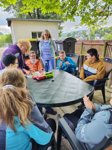 Gruppe von Kindern und Erwachsenen am Tisch im Freien, mit Spielen und lebhaften Gesprächen.
