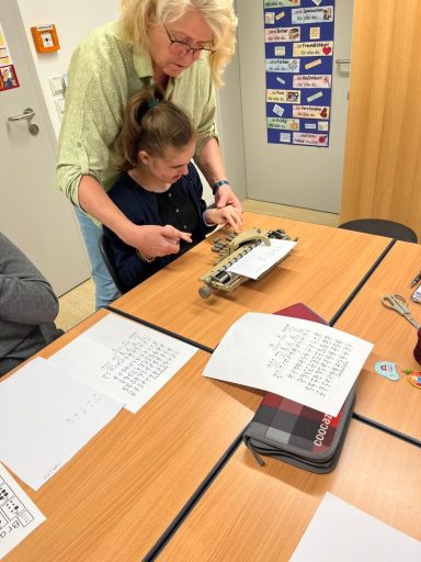 Eine Person hilft einer anderen, einen Brailledrucker zu bedienen. Unterrichtssituation.