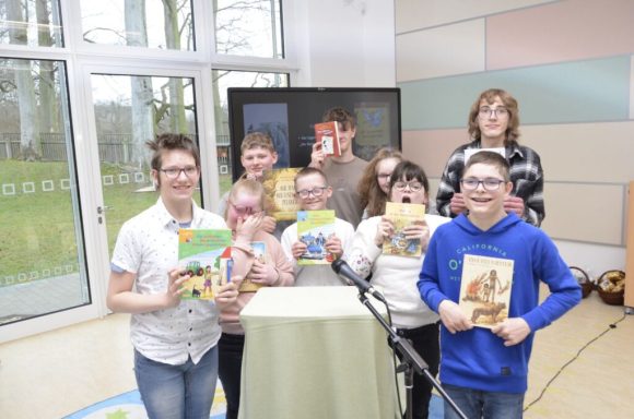 Gruppe von Kindern mit Büchern, lächelnd, vor einem Tisch in einem hellen Raum.