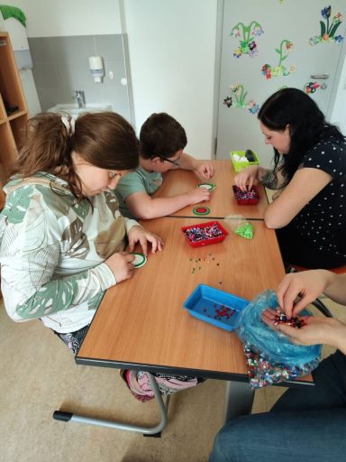 Drei Personen basteln an einem Tisch mit buntem Material in einem hellen Raum.