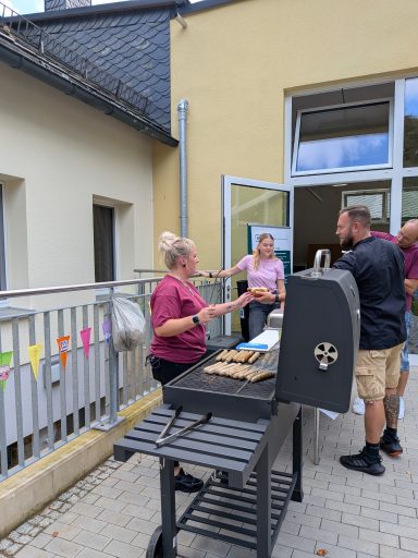 Frau am Grill mit Gästen auf einer Terrasse, fröhliche Atmosphäre.